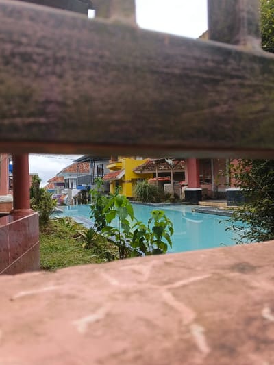 The image presents an outdoor scene in Pô Kô, Vietnam, viewed through two out-of-focus horizontal barriers. A dark, textured bar occupies the upper mid-ground, while a lighter, irregular stone-patterned barrier forms the lower foreground. The central feature is a calm, light blue swimming pool, displaying visible steps on its left side. Surrounding the pool is lush green vegetation, comprising manicured grass, various shrubs, and a prominent leafy green plant located between the foreground barrier and the pool. To the left of the pool, a reddish-brown pillar is visible, leading to a series of multi-story buildings with terracotta-tiled roofs and facades in white, grey, and purplish tones, appearing to ascend a gentle incline. To the right of the pool, a distinctive yellow building with a terracotta roof is visible, accompanied by a structure featuring reddish-pink pillars and a matching roof, surrounded by ornamental grasses and bushes. The background consists of additional buildings and an overcast, light grey sky. No individuals, actions, or interactions are present in the depicted scene.