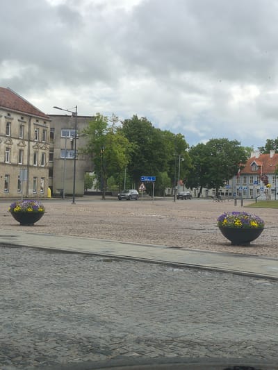 This is a medium shot, eye-level, outdoor, daytime photograph taken in the city of Šilutė, Lithuania. The image depicts a cobblestone square with two large planters filled with yellow and purple flowers. In the background, several buildings are visible, including a light-colored apartment building with an orange-tiled roof on the left and a gray building with a similar roof on the right. Trees are scattered throughout the scene, and a few cars are parked on the street. A blue road sign with white lettering is also visible, indicating directions to "ŠILALĖ" and "TAURAGĖ". The sky is overcast with clouds.