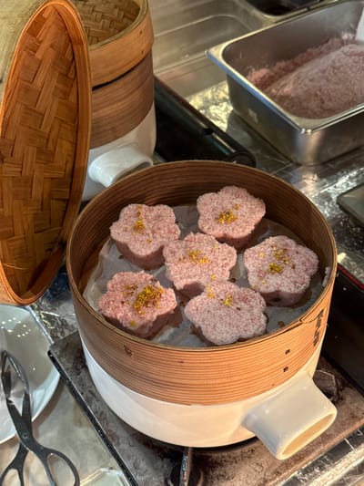 Six pink, flower-shaped steamed cakes are displayed within a light brown bamboo steamer, which rests on a white ceramic base with an integrated spout, on a metallic surface in Tongxiang, China. Each cake is topped with a light pink granulated texture and garnished with small golden-yellow decorative elements. The cakes are positioned on white parchment paper inside the steamer. To the immediate left, an open bamboo steamer lid leans against the main steamer. Behind the primary steamer, two additional stacked bamboo steamer baskets are partially visible. In the upper right background, a stainless steel pan holds a quantity of pink granulated material. A pair of silver-colored serving tongs with serrated edges is visible in the lower left, resting on a white plate or surface. No individuals are present in the scene.