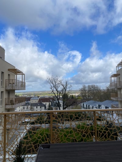 The image shows a view from a balcony in Saint-Cyr-l'École, France. The balcony features a decorative, patterned metal railing. In the distance, there are buildings and trees. The sky is partially cloudy.
