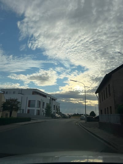 This media file depicts a street scene in Heinsberg, Germany. The camera is positioned at an angle, suggesting it's taken from inside a vehicle. The foreground is dominated by a dark, reflective surface, likely a car window. Beyond this, a street runs diagonally through the frame, bordered by modern white buildings on the left and a darker, older brick building on the right.

The sky is filled with dramatic clouds, with streaks of sunlight breaking through, indicating it is either sunrise or sunset. The lighting suggests it is late afternoon or early evening. Several parked cars are visible along the street, and there are streetlights present. The architecture of the modern buildings is characterized by clean lines and large windows, while the older building appears to be made of brick with rows of darker-colored windows. A palm tree is visible in front of one of the modern buildings, adding a touch of greenery to the scene.

There are no people visible in the frame. The overall mood is calm and serene, with the striking cloud formations and the golden light adding a visually appealing quality to the urban landscape. No text is visible in the image.