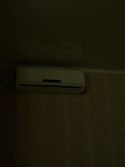 An air conditioner unit is mounted on a wall with ornate wallpaper. The air conditioner is white and has the brand name "Haier" visible on its front. A power cord extends from the unit and plugs into an outlet on the wall. The wallpaper is a dark color with a repeating damask pattern. The lighting in the room is dim, casting shadows and making the details of the wallpaper less distinct. The scene appears to be indoors, in a residential or commercial space. There are no people or animals visible in the image. There is another, less distinct, object mounted higher up on the wall, possibly another air conditioner unit, partially obscured by darkness. The location is specified as Zhovte, Ukraine.