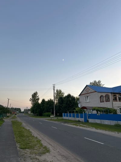 The image depicts a rural road in the evening. The sky is clear and light blue, with a crescent moon visible. A paved road stretches into the distance, flanked by a sidewalk and grassy verges. On the right side of the road, a two-story house with a blue roof and white brick exterior is visible, surrounded by a blue fence. Trees and power lines are also present in the scene, suggesting a semi-urban or village setting. There are no people visible in the image. The overall impression is one of a quiet, possibly residential area in a rural part of Belarus.