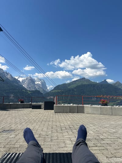 The image, taken on a sunny day in the city of Hasliberg Reuti, Switzerland, presents a scenic view from a terrace. In the foreground, a person's legs, clad in dark pants and blue socks, are extended, resting on what appears to be outdoor furniture. The terrace itself is paved with grey interlocking stones, and a low concrete wall with a red railing borders the edge, offering a view of the majestic Swiss Alps. In the background, snow-capped mountains rise majestically against a clear blue sky dotted with fluffy white clouds. The slopes of the mountains are covered with lush green trees, and some patches of snow are visible at higher elevations. Several wires, likely part of a cable car system, crisscross the sky. A few potted plants with pink flowers add a touch of color to the scene. The atmosphere is one of relaxation and appreciation of the natural beauty of the Swiss landscape.