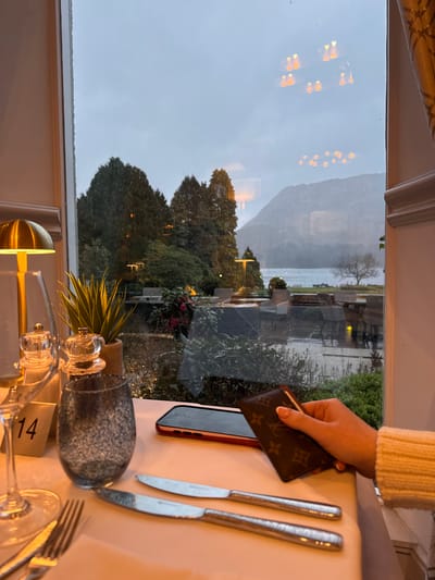 A table setting in Glenridding, United Kingdom, viewed through a window overlooking a lake and mountainous terrain, obscured by rain. On the table, there is a phone, a brown Louis Vuitton wallet being held by a hand wearing a white sweater, a fork and two knives, a wine glass with a visible table number '14', and a small potted plant on the window sill. The external environment is visible through the rain-covered window, featuring trees, a lake, and mountains under an overcast sky.