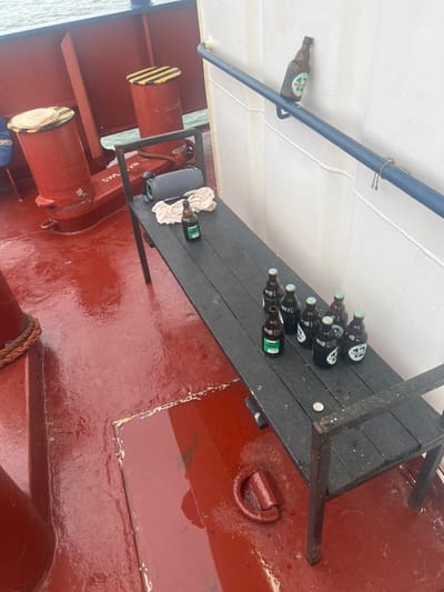 The image depicts a section of a vessel's deck, reportedly located in or around Bangor, United Kingdom. The scene is devoid of people.
The deck is painted a glossy red and appears wet, possibly from rain or sea spray. In the foreground and midground, a dark grey or black slatted bench with metal legs is positioned. On the left end of the bench, a crumpled light-colored cloth or towel rests next to a grey cylindrical object, possibly a portable speaker with a dark strap. A dark-colored beer bottle with a light cap and a green label ("HAWKSMOOR") stands upright on the bench near these items.
Further down the bench to the right, seven additional dark-colored beer bottles with light-colored caps are arranged. Several of these bottles also display visible "HAWKSMOOR" branding on their labels. One empty bottle cap lies on the bench surface.
Against a white vertical bulkhead to the right, a blue metal handrail runs horizontally. Balanced on this handrail is a brown beer bottle with a light-colored label prominently featuring "NANTLLE 44".
In the background and to the left of the bench, two large red cylindrical bollards are visible on the wet deck. The left bollard has a cream-colored top, possibly a covered capstan, while the right bollard features black and yellow warning stripes on its top. Ropes are visible coiled around the base of the left bollard. Beyond the vessel's structure, a body of water, appearing grey-green under an overcast sky, is visible. The deck also features a metal d-ring and a rectangular hatch cover.