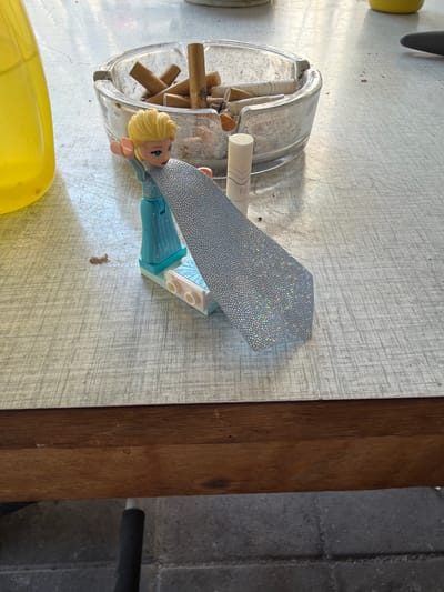 The image shows a close-up view of a plastic toy figure with blonde hair and a blue dress, resembling Elsa from Frozen, positioned on a table. The figure is holding a blue, sparkly fabric that resembles a cape. Next to the figure is a glass ashtray filled with cigarette butts. To the left of the ashtray is a bright yellow liquid in a clear container, possibly a bottle or jug. A white object with the letters "TE" visible is also near the ashtray and the toy. The table surface has a textured pattern. In the background, a wooden surface is visible below the table, and a tiled floor can be seen at the bottom of the image. The scene appears to be outdoors, likely on a patio or balcony, given the lighting and the presence of an ashtray. The question about the location in Boxtel, Netherlands, cannot be answered from the visual information provided in the image.