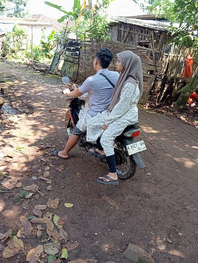 A male and female are depicted on a dark-colored motorcycle positioned on a reddish-brown dirt path in Bulukandang, Indonesia. The male driver, with short dark hair, wears a light grey short-sleeved shirt, dark shorts, and dark flip-flops. He is seated on the motorcycle with his hands on the handlebars. The female passenger, seated behind him, wears a light brown hijab, a long-sleeved white top with a faint pattern over dark leggings, and dark platform flip-flops. Her head is turned slightly to her right, looking forward. The motorcycle's rear visible license plate reads "W 6927 BH" with "03.26" below, indicating a registration validity. The ground is primarily dry dirt with scattered brown leaves, transitioning to a section of light-colored paving or brickwork under the vehicle and to the left. The background features lush green foliage, including banana plants, and distant light-colored residential buildings on the left. To the right, a dilapidated wooden structure, possibly a shed or animal pen constructed from slats and woven material, is partially visible amidst more vegetation; an orange object is near its base. The scene is illuminated by natural daylight.