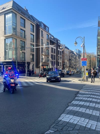 A motorcycle policeman, wearing a red helmet and blue and red jacket, is stopped at a crosswalk. The motorcycle is black and has a blue flashing light. The policeman is looking to his right. Behind the policeman, a black car with tinted windows is stopped at the crosswalk. The car has flashing blue lights on the roof. The car is parked on the right side of the street. To the left of the motorcycle, a street sign with the word "PAUL" is visible. The street is paved with grey asphalt and black and white checkered tiles. Several people are walking on the sidewalk behind the car. The buildings on the left side of the street are multi-story, with large windows and brick walls. The buildings are located in the city of Etterbeek, Belgium, at the coordinates 50.84 latitude, 4.382 longitude. The sky is clear and blue. The sun is shining. The air is clear. The time of day is daytime. The temperature is warm. The weather is pleasant.