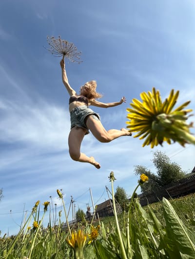 The image is a low-angle shot of a woman in mid-air against a bright, slightly cloudy blue sky. She is wearing a brown top and jean shorts. Her arms are extended, one holding a dried flower head. Below her are dandelions and other greenery, suggesting a field or meadow. In the background, there is a partial view of a wooden fence and a building.
The location is Усть-Тунтор, Russia.