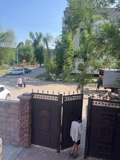 In a courtyard area of an apartment building in Aksu, Kazakhstan, two women stand near a parked white SUV and a dark green car. One woman wears a pink shirt and dark pants, while the other is in a white shirt and dark pants. A playground with colorful structures is visible in the background. Further back, a large box truck is being loaded by two individuals. One man in a patterned shirt and shorts is seen in the foreground of the truck, while another person is partially visible inside. Various cardboard boxes and other items are scattered around the truck. The scene is set under a clear blue sky with trees providing shade. In the foreground, a black metal gate with decorative elements is partially open, revealing a brick wall and a paved walkway.