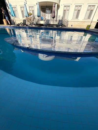 A person is standing on the edge of a swimming pool, holding a yellow pole with a bag attached to it. The person is wearing shorts and a hoodie. The swimming pool has blue tiles on the bottom and edges. The water in the pool is clear and reflects the surrounding environment, which includes a large building with windows and an umbrella. There are also chairs and tables visible around the pool area. The scene is set in Vaughan, Canada, likely a residential area given the presence of a pool and a large house. The overall impression is a sunny day at a home with a well-maintained pool.