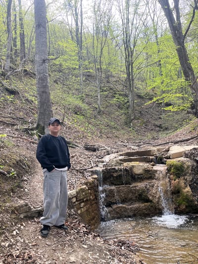 A person stands on a leaf-strewn ground next to a waterfall in a wooded area in Medven, Bulgaria. The individual wears a black sweatshirt, grey pants, and a cap. A small waterfall cascades over rocks and moss. Trees with new green leaves border the scene.