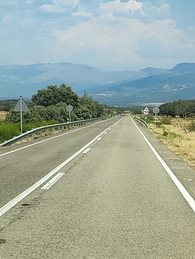 A straight asphalt road with white lane markings extends into the distance, bordered by a metal guardrail on the left and dry grass and trees on the right. In the background, a hazy mountain range under a cloudy sky is visible. Two triangular road signs are present: one on the left depicting a shield and another on the right with an image of a deer. The setting appears to be rural, consistent with the location of Zarza de Granadilla, Spain. There are no people or vehicles visible in the image.