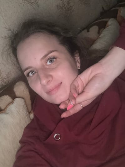 The media file is a close-up, over-the-shoulder selfie of a young woman lying down. She is wearing a dark red, long-sleeved garment with a metal grommet detail on the sleeve. Her hair is dark and appears to be loosely curled, falling around her face and shoulders. Her right hand is raised, partially covering her face, with manicured nails painted bright pink. She is wearing a ring on her ring finger.
The woman has light skin, light blue eyes, and is looking towards the camera. Her facial expression is neutral or slightly contemplative. The background consists of a textured surface, possibly a couch or bed, with a light beige and brown patterned fabric. The lighting is soft and appears to be indoor, suggesting it is either daytime or evening. The image is oriented sideways, as if taken while the subject is lying on their side. No text is visible in the image. The location context of Severnyy, Russia, does not provide any specific visual cues within the image itself.