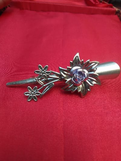 A silver-toned metallic decorative clip, possibly a hair barrette or brooch, is positioned diagonally on a red fabric surface. The clip measures approximately 9-10 cm in length. It features a prominent floral motif, with a central large flower composed of metallic petals surrounding a round, purple-hued, possibly jewel or enamel, center. This main flower is flanked by several metallic leaves. Extending leftward from the central design is a stem-like element adorned with three smaller, five-petal metallic flowers. The base of the clip, indicative of a barrette mechanism, is visible on the right end. The background consists of a smooth, deeply red textile, likely satin or silk, exhibiting minor folds and variations in sheen. The object is illuminated by a general light source, casting subtle reflections. This scene is located in Horažďovice, Czechia.