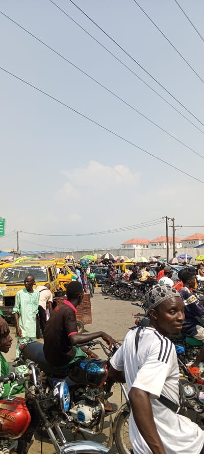 The image depicts a crowded outdoor street scene in Awjaw, Nigeria, characterized by significant pedestrian and vehicular traffic under a bright, partly cloudy sky. In the immediate foreground, multiple individuals are seated on motorcycles. One male subject on the far right wears a white short-sleeved shirt with black stripes on the sleeves and a patterned skullcap, looking towards the right. Another male subject is seated on a black motorcycle with blue detailing on the fuel tank, wearing a black t-shirt with red trim. A red motorcycle is partially visible on the far left.
The midground is densely populated with people and vehicles. Numerous yellow minibuses, common local transport, are visible, some appearing to be stationary. One minibus has "ALOMA" text on its side. Individuals are walking and standing between vehicles and stalls. A male figure in a light green traditional garment stands facing left, observing an off-frame area. Market activity is suggested by colorful vendor umbrellas providing shade. A green road sign on the far left indicates locations such as "ALABAR OWO" and "OKPE MKT. P.J.R."
In the background, additional people and vehicles are present. An electricity pole with multiple wires stands on the right. Beyond the immediate street level activity, a light grey wall or barrier is visible, topped by several modern residential buildings featuring orange-red pitched roofs and white facades. The overall environment suggests a bustling commercial or transportation hub.