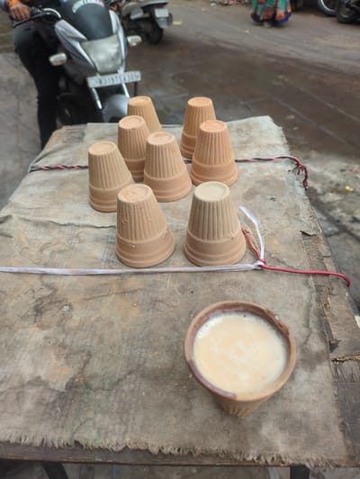 An outdoor street scene in Jodhpur, India, shows a wooden cart with a dirty, greyish-brown cloth covering. Arranged on the cart are seven small, unglazed clay cups, stacked in two rows. In the foreground, a single clay cup is filled with a milky, light-brown liquid, likely chai. Behind the cart, a motorcycle with a license plate is partially visible, along with the legs of a person in dark pants. In the background, a blurry figure in a colorful skirt walks away, and other motorcycles are parked. The ground appears wet, suggesting recent rain or a cleaning.