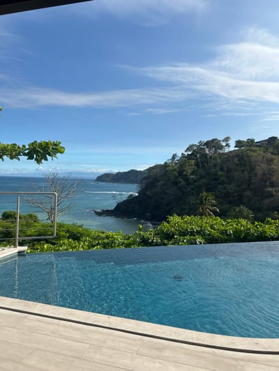The image depicts a swimming pool with a view of the Pacific Ocean and a forested hillside, taken from the edge of a private residence in Playa Herradura, Costa Rica. The pool is rectangular, surrounded by light-colored tiles, and the water is a clear, deep blue. The hillside behind the pool is covered in lush vegetation, with a rocky outcropping visible in the distance. The sky is a bright blue with thin, white clouds. The ocean is calm and the water is a deep turquoise. The image captures a serene, tropical setting, suggesting luxury and relaxation. The location is a resort area, providing a view of the ocean. There are no people in the image. The image suggests a luxurious, tropical escape.