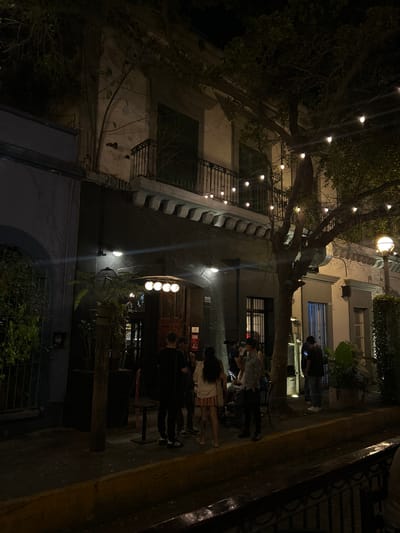 The image shows a nighttime scene in Mazatlán, Mexico. The street is dimly lit by overhead string lights and building-mounted fixtures. A multi-story building with a balcony and decorative facade is visible on the left. A tree with bare branches partially obscures the view of the building. On the right, a group of people is gathered outside what appears to be a bar or restaurant. Some individuals are standing, seemingly waiting to enter. The building is illuminated by several lights above the entrance. The sidewalk is narrow and lined with a curb. The overall ambiance suggests a lively nightlife scene.