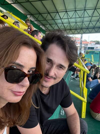 A close-up, slightly angled selfie shows a couple in the foreground, seated on stadium bleachers. The woman on the left, with reddish-brown hair and large, dark sunglasses, is looking directly at the camera. The man on the right, with dark, wavy hair and a mustache, is also looking towards the camera, with his arm resting on the seat. They are both wearing casual dark clothing.

In the background, other spectators are visible sitting on the green and yellow bleachers under a metal and translucent roof structure. Some individuals are standing near the railing, and a few are walking in the distance. The setting appears to be an outdoor sports venue. The architectural style of the buildings visible beyond the stadium suggests a European location. Based on the presence of the "Pingo Doce" supermarket chain logo visible on the stadium signage in the background, the location is likely in Portugal. Specifically, the stadium is identified as Complexo Desportivo de Santa Maria da Feira, located in the city of Santa Maria da Feira, Portugal.
