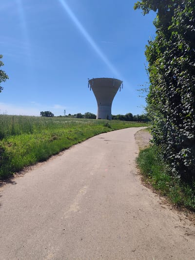 The image depicts a concrete water tower in Chaponnay, France. The tower is the central focal point, towering over a path leading towards it. The path is made of concrete and is bordered by green vegetation, including tall grasses and leafy trees and bushes. The sky is clear and blue, with a sun glare visible in the top portion of the image. A faint contrail from an aircraft crosses the sky above the tower. A small utility tower is visible between the water tower and tree line. The scene suggests a rural or semi-rural environment.