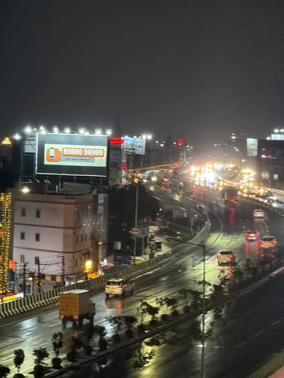 The image depicts a nighttime scene on a wet, multi-lane highway in Shamshabad, India, likely during or shortly after rainfall, evidenced by the reflections on the road surface. The atmosphere is dark and hazy, with numerous vehicle headlights and streetlights creating streaks of light and illuminating the wet asphalt.
In the foreground, a white box truck with a wooden cargo container is traveling on the right lane of a lower highway. Further up and to the left, a white SUV is also on the same lane. A multi-story building with white walls and dark windows is visible on the left, adorned with a string of yellow lights. A billboard above the building displays an advertisement with the number "89880 96999" and "FOR ADS CONTACT" in orange text.
On the upper highway, several vehicles are visible, their headlights and taillights creating a blurred effect due to the low light conditions and motion. The highway infrastructure includes elevated sections and concrete barriers. In the background, the city skyline is dimly visible, with various lights indicating urban development. The overall scene conveys a busy, urban environment at night, with traffic flow on wet roads.