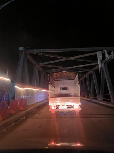 The image presents a nighttime view from a vehicle, looking forward at a white truck on a metal truss bridge in Lhoknga, Indonesia. The dark sky looms above the bridge's skeletal structure of crisscrossing beams.
A white, box-body truck, identified by "ISUZU" on its rear, is the primary subject in the midground, its red taillights glowing. A license plate displaying "BL 8292-C" is visible on the truck's rear. The truck's cargo bed is covered by a dark tarp, on which partial text can be seen, appearing to read "MUSYA..." and "...AH DE..." or "...ANH DE...".
To the left of the roadway, a temporary barrier system is erected. It features orange plastic barriers at the base and a taller, white, semi-translucent partition. A string of bright, elongated yellow-orange lights runs horizontally along the top of this temporary wall, creating light streaks due to exposure or motion. A faint human figure is discernible behind these lights on the far left. The road surface appears somewhat damp or reflective, with scattered debris along the edge of the pedestrian path. Reflections from the truck's lights and other ambient light sources are visible on the road and in the immediate foreground, likely from the dashboard or windshield of the viewer's vehicle. The scene suggests travel at night, possibly encountering roadwork or a temporary setup on the bridge.