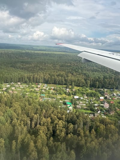 An aerial view captures a rural settlement nestled amongst dense, green forests, with an airplane wing visible in the upper right quadrant. The settlement, appearing to be in the vicinity of Vozhoi, Russia, is characterized by numerous small houses with distinct roofs, surrounded by gardens and small plots of land. The terrain slopes gently, with patches of lighter green suggesting open fields or cultivated areas interspersed with the darker, more varied shades of the forest canopy. The sky above is partially covered by dynamic clouds, with hints of blue sky visible. The image provides a snapshot of a semi-rural landscape from an elevated perspective, likely during daytime, under mixed weather conditions.