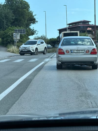 The image captures a street scene in Vrijesno, Montenegro, likely on a cloudy day. In the foreground, a silver Lexus GS 300 is visible from the rear, displaying a license plate reading "AI 7379 TT". The car has minor damage to its lower rear bumper. Ahead of the Lexus, a white Toyota RAV4 is stopped at a pedestrian crossing. The RAV4's license plate is partially obscured but shows "E 580 CE 69". In the background, a bus and a building with a distinctive wooden architectural style and a red roof are present. Traffic signs and streetlights are also visible, indicating an urban or suburban environment. The road surface is asphalt, and white lines delineate the pedestrian crossing and traffic lanes. The overall scene suggests normal daytime traffic conditions.