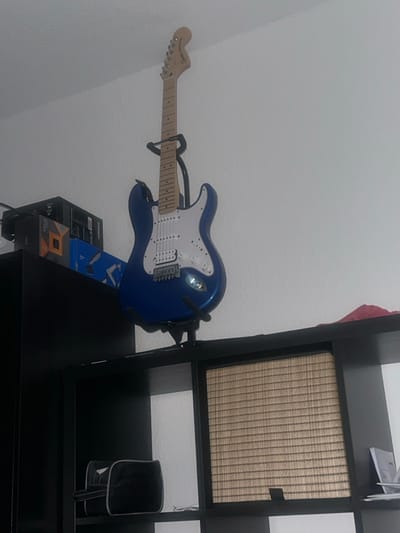 The image displays an indoor scene in Nordhausen, Germany. A blue Squier by Fender electric guitar, featuring a white pickguard, light wooden neck, and chrome hardware, is prominently positioned upright on a black stand. The stand rests on the top surface of a black, cube-style shelving unit.
To the immediate left of the guitar, on the top surface of the same black shelving unit, a stack of cardboard boxes is visible, including a dark box, one with an orange/gold square logo, and a blue box. Wires are also present in this area.
Below the guitar, the black shelving unit has visible compartments. The leftmost compartment contains a black soft-sided case with grey trim. The central compartment is fitted with a light brown, horizontally slatted or woven insert. The rightmost compartment holds a stack of white papers or documents. A small, crumpled red fabric object is partially visible on the top right surface of the shelving unit. The walls and ceiling are uniformly white and appear to have a textured finish. No individuals are present in the scene.
