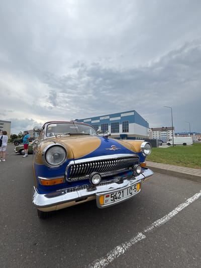In the city of Uzda, Belarus, an aged, two-toned car, yellow and blue, is parked on asphalt. The car's chrome grille and circular headlights gleam under an overcast sky. A license plate displaying "BY 5421 IC-5" is affixed to the front. In the background, a modern building with blue and white accents stands, along with residential buildings and a white truck. Several individuals are visible near other vehicles, one wearing a blue shirt and red shorts, and another a white shirt and shorts. The scene suggests an outdoor event or gathering focused on vintage automobiles.