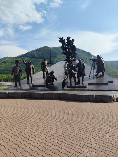 This media file captures a bronze monument depicting several figures engaged in various activities. The monument is situated outdoors with a mountainous, forested landscape serving as a backdrop. The sky is bright blue with scattered white clouds, suggesting a pleasant day, likely midday due to the strong shadows cast by the figures and the overall bright lighting.
The monument itself is built on a dark, polished stone base and features several life-sized bronze statues. In the foreground, from left to right, are figures appearing to be workers or explorers. One stands tall with his arm raised. To his right, another figure kneels, examining something on the ground. Further to the right, another group of figures stands on a higher platform. One is operating a surveying instrument on a tripod, while two others appear to be consulting a map or blueprint. A central abstract bronze sculpture rises above these figures, adding a dramatic element to the composition.
The setting appears to be a public space, possibly a park or memorial site, paved with reddish interlocking stones in the foreground. In the background, a wide, flat river or body of water is visible, with a bridge or structure crossing it on the left. The foothills of a large mountain range, densely covered with green trees, dominate the background. The presence of a large dam structure can be seen in the far distance on the right.
While the specific event or historical context is not explicitly detailed, the monument's themes of exploration, surveying, and perhaps construction are evident through the poses and objects depicted. The overall impression is one of historical significance and human endeavor in a natural, rugged environment. The location context provided (Cheremushki, Russia) suggests the monument might commemorate a significant event or individuals related to the development or exploration of this region.
No visible text is clearly decipherable in the provided media.