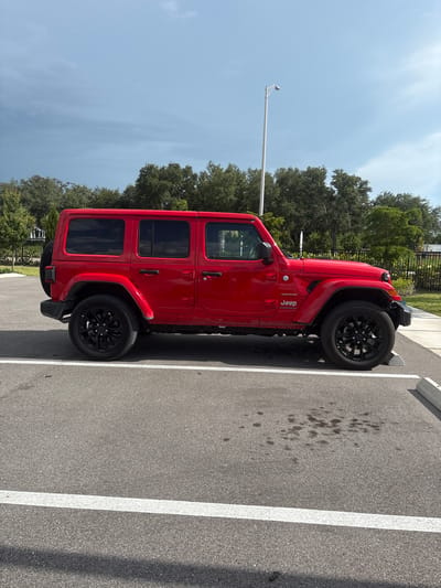 A red Jeep Wrangler Sahara is parked in a lot with a light grey sky. The vehicle is parked in a marked parking space with white lines. The Jeep is a four-door model with black rims. Behind the Jeep, there is a row of trees and a black metal fence. In the distance, there are buildings and a street lamp. The ground is asphalt with a few dark stains. The image appears to be taken in Sarasota, Florida, United States.