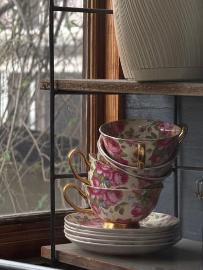 The image shows a stack of four vintage teacups with a floral pattern, featuring pink roses and blue accents, on a wooden shelf. The cups have gold handles and rims. Below the cups, a stack of saucers with the same floral pattern is visible, topped by a white saucer. Behind the teacups and saucers, a window with bare branches of trees is visible, suggesting an outdoor scene. The shelf is part of a multi-tiered shelving unit, with another shelf above it holding a ribbed, beige planter. The overall scene evokes a rustic, vintage aesthetic. The location is not specified in the image.