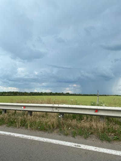The image captures a view from a roadside in Kislyakovskaya, Russia, looking across a green field under a dramatic, overcast sky. A metal guardrail runs horizontally across the foreground, separating the road from the grassy shoulder and the field beyond. The guardrail has red reflectors on it. In the mid-ground, a band of trees forms a distant treeline. To the right of the frame, a tall pole with some equipment on top stands in the field. A distinct rain shower is visible in the distance, indicated by a dark vertical streak against the sky. The overall atmosphere is one of impending or ongoing weather change. There are no people or animals visible in the image.