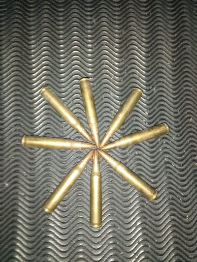 Ten unfired rifle cartridges are arranged in a precise radial pattern, forming a ten-pointed star with all projectiles converging at a central point. Each cartridge features a brass-colored casing and a pointed, copper-jacketed projectile. They rest on a dark, monochromatic surface characterized by a repetitive, wavy, ridged texture, consistent with a heavy-duty rubber or plastic mat. The lighting is uniform, highlighting the metallic finish of the cartridges. No individuals or discernible actions are present. The specific location within the city of Tas-Kop', Kazakhstan, is not identifiable from the visual data provided.