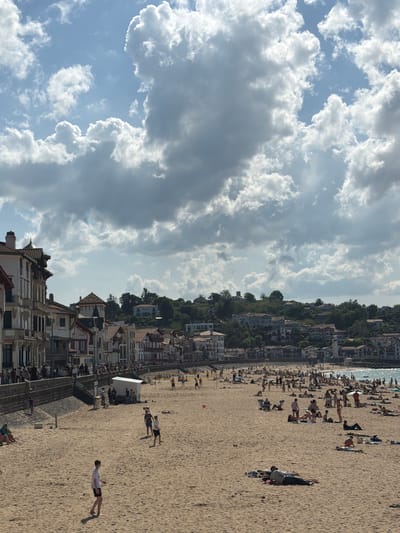 The image captures a beach scene in Saint-Jean-de-Luz, France. A sandy beach is populated with numerous people sunbathing, walking, and interacting in small groups. The beach is bordered by a line of buildings, including houses and structures with distinctive Basque architecture, characterized by white walls and red or brown tiled roofs. In the background, there is a hillside with more buildings and trees. The sky is filled with cumulus clouds.