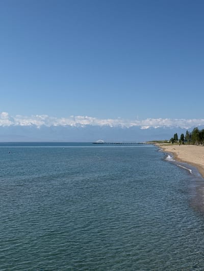 The image shows a tranquil scene at the beach in Koshkol', Kyrgyzstan. A vast expanse of calm, blue water stretches towards a distant shore lined with a sandy beach. In the background, majestic snow-capped mountains form a dramatic horizon under a clear, bright blue sky. A long pier extends from the shore into the water, with a small building or structure at its end. Trees dot the shoreline, adding a touch of green to the landscape. The beach itself appears mostly empty, with only a few buoys visible in the water, indicating a peaceful and serene atmosphere. The light suggests it is daytime, with the sun casting gentle reflections on the water's surface.