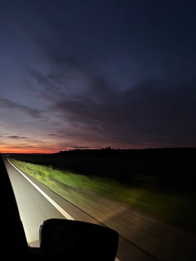 The image depicts a view from inside a vehicle driving on a road at dusk. The sky displays a gradient of colors, transitioning from a deep blue at the top to orange and pink hues near the horizon. Dark clouds are scattered across the sky. The road is paved with white lines marking the lanes, and the headlights of the vehicle illuminate the road and the blurred green vegetation to the side. A dark silhouette of trees and a treeline is visible in the distance. The bottom portion of the image shows a dark shape, likely a side mirror or part of the car's interior. The image does not contain any identifiable people or specific landmarks that would indicate the location in Izh-Boyki, Russia.