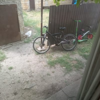 The image depicts an outdoor scene in Slobidka, Ukraine, likely a residential or backyard area. In the foreground, a black mountain bike with front suspension and disk brakes is parked on cracked, dry earth with sparse grass. Behind the bicycle, a brown wooden fence serves as a backdrop. To the right of the mountain bike, a green scooter with silver handlebars stands upright. Further to the right and slightly behind the scooter, a small child's bicycle with a silver frame and red wheels is visible. To the left of the mountain bike, a portion of a weathered, dark brown door or gate set into a stone structure is present. A crumpled white plastic bag is on the ground near the stone structure. The environment appears somewhat unkempt, with areas of dry earth and patchy grass. The lighting suggests it is daytime, possibly overcast.