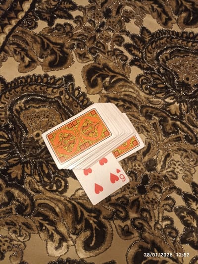 The image displays a deck of playing cards placed on an intricately patterned fabric. The fabric, which serves as the background, features a rich design with large, swirling paisley-like motifs in shades of brown and black, accented with shimmering gold or silver glitter threads against a beige base.
A stack of playing cards lies on the fabric, oriented diagonally from the upper left towards the lower right. The visible top card of the deck shows its back, which has an ornate, symmetrical design in orange and gold. Partially peeking out from beneath the main deck is a single playing card, revealing its face. This card is clearly identifiable as the 9 of Hearts, distinguished by its nine red heart pips and the numeral "9" at one corner.
The scene appears to be an indoor setting, with soft, possibly artificial, lighting highlighting the glitter in the fabric. No human subjects are present, and no specific activity is being performed with the cards; they are simply arranged on the surface. The provided location context is Khidirsho, Uzbekistan.
A digital timestamp is visible in the bottom right corner of the image, reading "28/01/2026 12:52". This indicates the date and time the photograph was taken, suggesting a midday capture. The overall impression is one of a casual, quiet moment, perhaps a pause during a card game or a decorative setup.