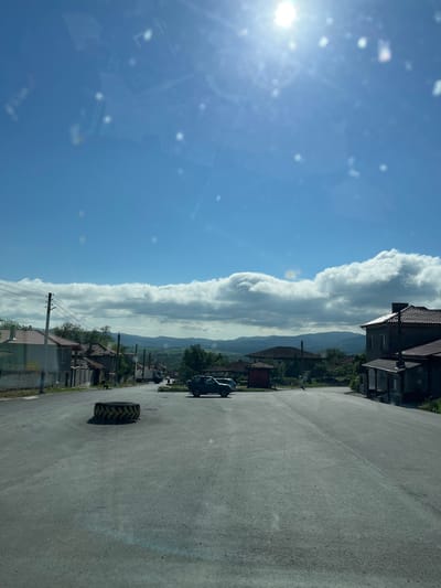 A sunny day in Rish, Bulgaria. The image is taken from inside a vehicle, looking out onto a street. A large tire is in the middle of the road. Houses line one side of the street, with power lines visible. Mountains are visible in the distance behind the houses. Cumulus clouds are on the horizon. The sky is blue with a bright sun on the left.