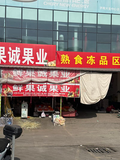 The image depicts an outdoor commercial scene in Licheng District, Jinan, China. In the background, a multi-story building with a reflective, grid-patterned glass facade displays a banner featuring "CHERY NEW ENERGY" and "济南大金中心店" (Jinan Dajin Center Store).

Below the building, a fruit market and an adjacent shop are visible under bright sunlight. The fruit market entrance is marked by two prominent red banners: a larger one reading "诚果业" (Cheng Guoye - Honest Fruit Industry) and a smaller one below it stating "鲜果诚果业" (Xianguo Chengguoye - Fresh Fruit Honest Fruit Industry), adorned with fruit illustrations and contact numbers. To the right, another red banner indicates a "熟食冻品区" (Shushi Dongpin Qu - Cooked Food Frozen Products Area), next to an open storefront partially obscured by a rolled-back cream-colored tarp.

Inside the fruit market, bundles of sugarcane stalks are stacked vertically on the left, adjacent to a silver-toned sugarcane juicer machine. Piles of discarded sugarcane pulp are scattered on the ground directly in front of the machine. Various fresh fruits, including oranges and pineapples, are displayed on tables and in crates within the shop's entry area.

Inside the adjacent, dimly lit shop area to the right, a person wearing a red shirt is crouched low on the ground, near a small, light-colored dog.

The immediate foreground consists of a dark asphalt pavement. A rear section of a black scooter or motorcycle is visible on the far left. A white stand holding clear plastic bags containing packaged goods is positioned on the pavement near the fruit market entrance. A metal grate drain is embedded in the pavement on the right side of the frame. Strong shadows are cast across the ground, indicating direct sunlight from the upper left.