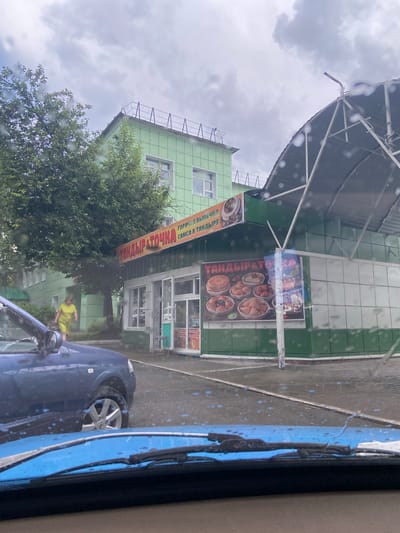 The image shows a street scene in Beloretsk, Russia, under a cloudy sky with raindrops on the camera lens. In the foreground, the blue hood and windshield wipers of a car are visible. To the right, a blue car is parked. Behind the car is a small food establishment with a green facade and a large awning. A sign above the entrance reads "ТАНДЫР & ТОЧКА" (Tandyr & Tochka), indicating a tandoor bakery. A woman in a yellow dress walks past the building. The building behind the food establishment is light green with multiple windows. The street is wet, suggesting it has been raining.