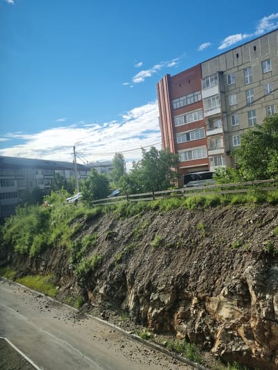 This image depicts a steep, rocky embankment with a road running along its base. The embankment is partially covered with green vegetation. At the top of the embankment, a wooden fence runs horizontally, with trees and a portion of a multi-story building visible behind it. The building appears to be made of brick and concrete, with multiple windows. Another, older-looking multi-story building is visible further in the background, along with a street light and power lines. The sky is blue with scattered white clouds. The location is identified as the city of Rezh, Russia.