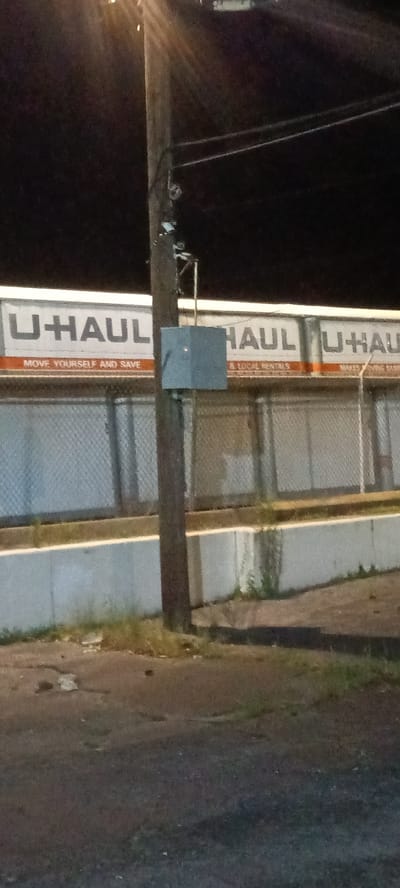 The image depicts a U-Haul facility at night in Fort Smith, Arkansas, United States. The facility is identifiable by the large "U-HAUL" signage on the building. A wooden utility pole is prominently positioned in the foreground, featuring a square metal box attached to it. Below the U-Haul signage is a concrete retaining wall, and there is a paved surface with some vegetation visible. The overall lighting is dim, suggesting it is late evening or night.