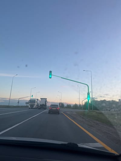 The image shows a view from inside a vehicle driving on a highway towards a traffic light in Kivsertkasy, Russia. The sky is a clear blue with some wispy clouds. The road has two lanes in the direction of travel, separated by a white line. A yellow line marks the right edge of the road. There are streetlights along the side of the road.
In the foreground, the windshield of the vehicle is visible, with some smudges and scratches. In the mid-ground, there are several vehicles on the road. Ahead, there are two white trucks, one of which appears to be a tanker truck. To the right of the trucks, a dark-colored car is visible. A traffic light in the distance is green, indicating that traffic can proceed. A speed limit sign displaying "75" is visible on the traffic light pole.
In the background, there are trees and a faint treeline. The overall scene depicts a typical highway intersection during what appears to be late afternoon or early evening, given the lighting conditions.