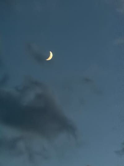 A crescent moon is visible in a dark blue sky, partially obscured by clouds. The moon is a bright yellow and is positioned in the upper right quadrant of the image. Dark gray clouds are scattered across the sky, with some appearing more dense than others. The overall mood of the image is serene and slightly mysterious. The location of Damianitsa, Bulgaria is not depicted in this image.