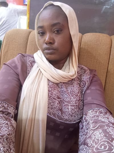 A person, presumably a woman, is seated indoors in a tan-colored, upholstered chair. She is wearing a mauve-colored, long-sleeved garment adorned with white decorative patterns and embellishments around the collar and cuffs. A cream-colored headscarf frames her face and drapes across her chest. The background is somewhat blurry, but another person wearing white is visible to the upper left. The location is purportedly Ajiwa, Nigeria, though visual elements confirming this are not present.